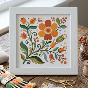 May include: A square white frame containing a cross-stitch embroidery on white fabric. The design features a large central orange flower surrounded by smaller orange and yellow tulips, green leaves, and red berries, all arranged in a symmetrical floral pattern.