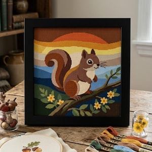 May include: A framed cross-stitch embroidery featuring a brown squirrel perched on a tree branch with yellow flowers. The background displays a layered sunset in shades of orange, yellow, and blue. The artwork is enclosed in a black square frame.