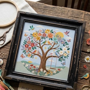 Könnte beinhalten: Ein quadratisches Kreuzstich-Stickbild in einem schwarzen Holzrahmen mit Gebrauchsspuren, das einen Baum mit bunten Blumen, verschiedenen Vögeln auf den Ästen, einer hängenden Schaukel, Schmetterlingen und einer gelben Sonne vor hellblauem Himmel zeigt.