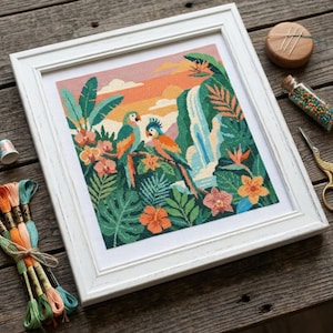 May include: Framed cross-stitch artwork featuring two parrots perched in a tropical jungle scene. The artwork includes a waterfall, vibrant flowers, and lush greenery, set against a sunset sky. The white frame complements the colorful design.