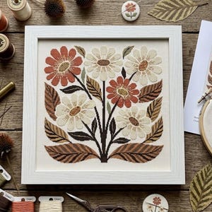 May include: A framed cross-stitch embroidery featuring a floral arrangement with cream, terracotta, and dark brown flowers and leaves. The design is centred on a white fabric background within a square white frame, showcasing detailed needlework.