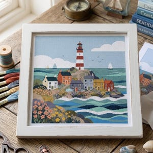 May include: A framed cross-stitch embroidery depicting a coastal scene with a red and white lighthouse, a cluster of small houses in various colours, and a rocky shoreline with flowers, all set against a blue sky and a wavy sea with sailboats.