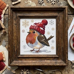 May include: A cross-stitch embroidery of a robin wearing a red knit hat with a pom-pom, surrounded by small embroidered snowflakes on a white fabric background, all encased within a rustic, dark brown wooden square frame with a textured finish.