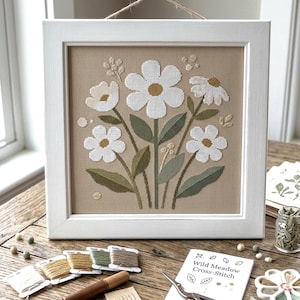 Puede incluir: Una obra de punto de cruz enmarcada que muestra un ramo de flores blancas con centros amarillos y hojas verdes sobre un fondo de tela color canela. El marco cuadrado de madera está pintado de blanco e incluye una cuerda de yute para colgar.