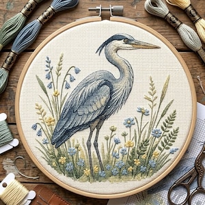 May include: A completed cross-stitch embroidery of a blue heron standing in a field of blue and yellow flowers and green grass. The heron is in shades of blue and gray. The embroidery is in a wooden hoop, with spools of thread around it.