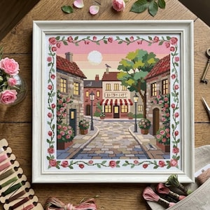 May include: A framed cross-stitch embroidery depicting a European street scene with stone buildings, a cafe with a red and white striped awning, a cobblestone path, and a border of pink roses, all set against a soft pink and yellow sunset sky.