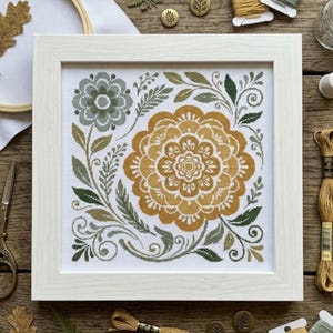 Könnte beinhalten: Ein quadratischer weißer Rahmen mit einer Kreuzstichstickerei auf weißem Stoff. Das Design zeigt ein großes, goldgelbes florales Mandala in der Mitte, umgeben von kleineren salbeigrünen Blüten, filigranen Blattranken und feinen Schnörkeln.