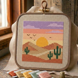 May include: A cross-stitch landscape scene featuring a desert sunset with cacti, mountains, and birds. The artwork is framed in a square wooden hoop with a leatherette hanger. Several spools of embroidery floss are visible.