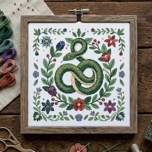 Könnte beinhalten: Kreuzstich-Stickerei mit einer grünen Schlange in einem floralen Kranz. Die Schlange hat einen hellbraunen Bauch, umgeben von grünen Blättern, roten, blauen und lila Blumen. Das Kunstwerk ist in einem quadratischen Holzrahmen gerahmt.