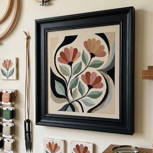 May include: A square framed cross-stitch embroidery featuring three stylised flowers with muted red petals and sage green leaves on a beige fabric background, enclosed in a black wooden frame with a decorative beveled edge and a neutral matting.