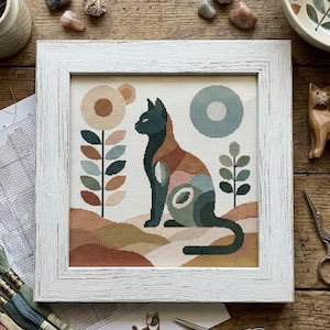 May include: A framed cross-stitch embroidery featuring a stylised cat in shades of dark green, brown, and muted pink, sitting on a landscape of rolling hills. The design includes a tall flower with circular petals and a circular sun in the sky.