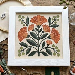 Puede incluir: Un marco cuadrado blanco que contiene un bordado de punto de cruz sobre tela beige. El diseño presenta un patrón floral simétrico con grandes pétalos naranjas en forma de abanico, hojas verde oscuro y pequeñas bayas verde claro.