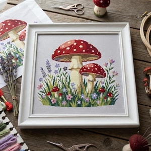 May include: A square white frame containing a cross-stitch embroidery of two large red-capped mushrooms with white spots, surrounded by smaller mushrooms, lavender sprigs, and various small blue, pink, and purple wildflowers on a light gray fabric.