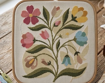 May include: A square wooden embroidery hoop containing a piece of white Aida fabric with a cross-stitch design of a central plant featuring various flowers in shades of pink, yellow, and blue, surrounded by dark green leaves and stems.