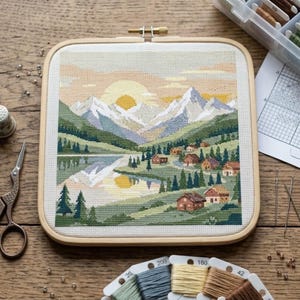 May include: Cross-stitch landscape scene featuring a mountain range, lake, and small village, framed in a square wooden hoop. The color palette includes greens, blues, and yellows, with a setting sun. Craft supplies, including thread and scissors, are visible.