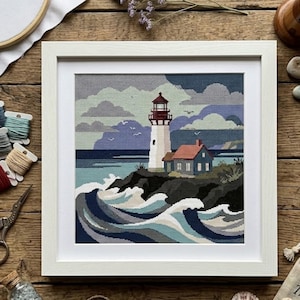 Könnte beinhalten: Eine gerahmte Kreuzstichstickerei, die einen weißen Leuchtturm mit rotem Dach und ein kleines angrenzendes Haus auf einer felsigen Klippe zeigt. Die Szene umfasst brechende blau-weiße Meereswellen, ein dunkles Meer und einen bewölkten Himmel.