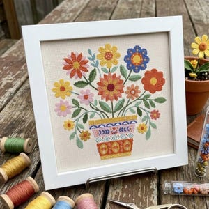 Peut inclure: Une broderie au point de croix encadrée représentant un bouquet de fleurs colorées dans un pot à motifs. Les fleurs sont réalisées dans des tons de jaune, orange, rose et bleu avec des feuilles vertes. L'ensemble est dans un cadre carré blanc.