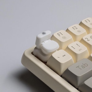 May include: A novelty white toilet-shaped keycap installed on a beige mechanical keyboard. The keycap replaces the F1 key. Other keys include numbers, symbols, and the Tab key.