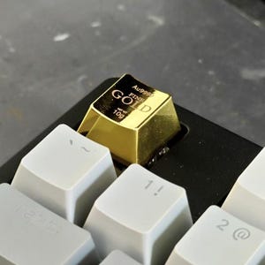 Könnte beinhalten: Ein goldfarbener Keycap in Form eines Goldbarrens, beschriftet mit "GOLD FINE" und "10g", sitzt auf einer weißen mechanischen Tastatur. Der Keycap ist von anderen weißen Tasten mit schwarzer Beschriftung umgeben. Die Tastatur befindet sich auf einer dunklen Oberfläche.