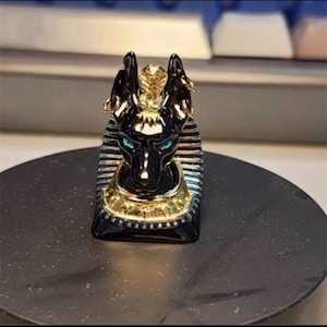 May include: A black and gold Anubis figurine with turquoise eyes. The figurine has a detailed design, including a cobra on the head and blue and gold accents. The figurine is on a dark surface.