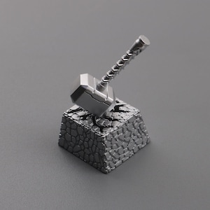 May include: A silver-colored miniature hammer, resting on a textured, gray stone base. The hammer's head is a rectangular shape, and the handle is detailed with a ridged design. The base has a cracked, stone-like appearance.