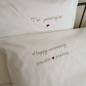 Puede incluir: Dos fundas de almohada blancas con texto bordado y pequeños corazones rojos. La funda superior dice "Ten years of us" y la inferior "Happy anniversary" seguida de las fechas "3.06.2016" y "3.06.2026" separadas por un corazón.
