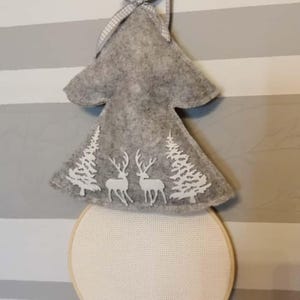 Może przedstawiać: Szara filcowa ozdoba choinkowa z białymi wycięciami w kształcie jeleni i drzew. Ozdoba ma okrągłą, jasną podstawę z tkaniny i kratowaną wstążkę do zawieszenia. Świąteczna dekoracja.