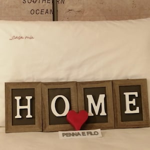 May include: A white pillow with the embroidered text "Casa mia" in red. Below, the word "HOME" is spelled out in individual framed letters, with a red heart between the "M" and "E". The text "PENNA E FILO" is below the heart.