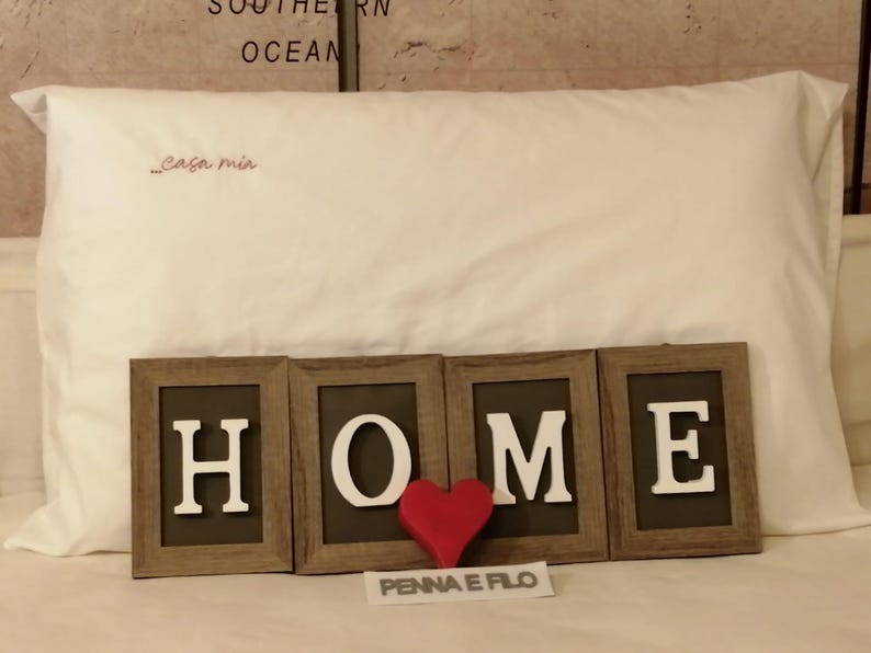 Op de afbeelding: Een wit kussen met de geborduurde tekst "Casa mia" in het rood. Daaronder staat het woord "HOME" in afzonderlijke ingelijste letters, met een rood hart tussen de "M" en de "E". De tekst "PENNA E FILO" staat onder het hart.