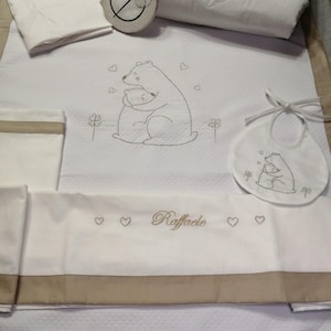 May include: A white baby bedding set with a bear and cub design, hearts, and the name "Raffaele" embroidered in gold. Includes a quilt, bib, and pillow. The set has a beige border.