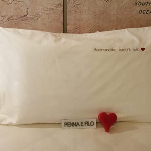 Può includere: Una federa rettangolare bianca con il testo italiano ricamato "Buonanotte amore mio" seguito da un piccolo cuore rosso. Un piccolo cartello rettangolare bianco con il testo nero "PENNA E FILO" e un oggetto a forma di cuore rosso sono davanti.