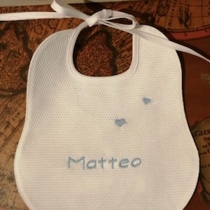 May include: White baby bib with the name "Matteo" embroidered in light blue. Two small blue hearts are embroidered above the name. The bib has a textured surface and a white ribbon tie.