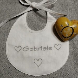 May include: White baby bib with the name "Gabriele" embroidered in gray, along with three gray heart designs. The bib has a tie closure and is next to a yellow heart-shaped stone.