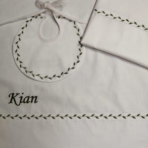 Puede incluir: Juego de babero y manta para bebé blancos. El babero tiene un diseño de hojas bordado alrededor del borde y un diseño de hojas a juego en la manta. El nombre "Kian" está bordado en la manta en color verde oscuro.