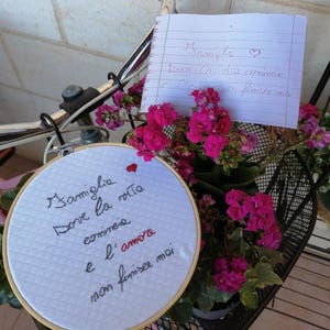 Könnte beinhalten: Ein Fahrradkorb mit leuchtend rosa Blüten und einem bestickten Stickrahmen. Der Rahmen trägt die Inschrift "Familie, wo das Leben beginnt und die Liebe nie endet". Eine handschriftliche Notiz ist ebenfalls zu sehen.