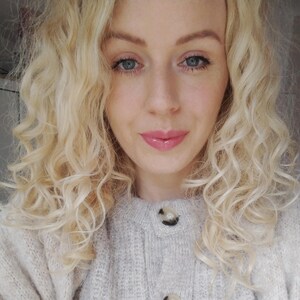 May include: A woman with long, blonde, curly hair wearing a light gray cardigan with two buttons.