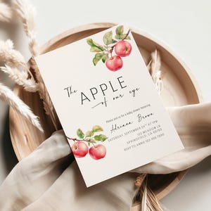 May include: Baby shower invitation with the text "The Apple of our eye". The invitation features watercolor illustrations of red apples and green leaves. The invitation is on a cream-colored card.