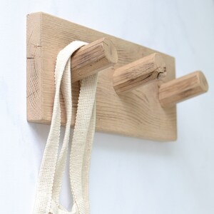 May include: A wooden wall-mounted coat rack with three hooks. The hooks are made from natural wood and have a rustic look. A white canvas tote bag is hanging on one of the hooks.