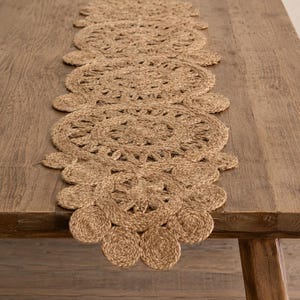 May include: A long, narrow table runner crafted from natural tan jute fiber. The runner features a repeating pattern of large, circular, braided medallions connected by smaller, solid circular motifs, creating a scalloped edge along the length.