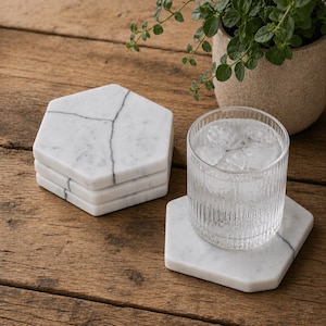 May include: A stack of three white hexagonal marble coasters with grey veining and one matching coaster supporting a clear, ribbed glass filled with ice and water. The coasters feature smooth, polished surfaces and distinct geometric shapes.