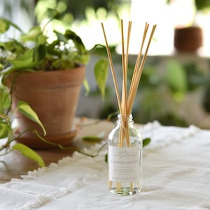 May include: A clear glass reed diffuser bottle with wooden reeds. The label reads "Garden House" and "Natural Green, Mint, Refreshing, Citrus". The bottle sits on a white tablecloth, with a potted plant in the background.