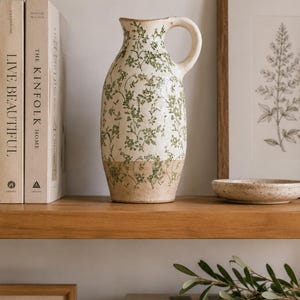 May include: A ceramic pitcher featuring a cream-colored base with a green floral vine pattern. The lower portion of the pitcher has a natural tan finish. It includes a curved handle on the right side and a slightly flared rim at the top opening.