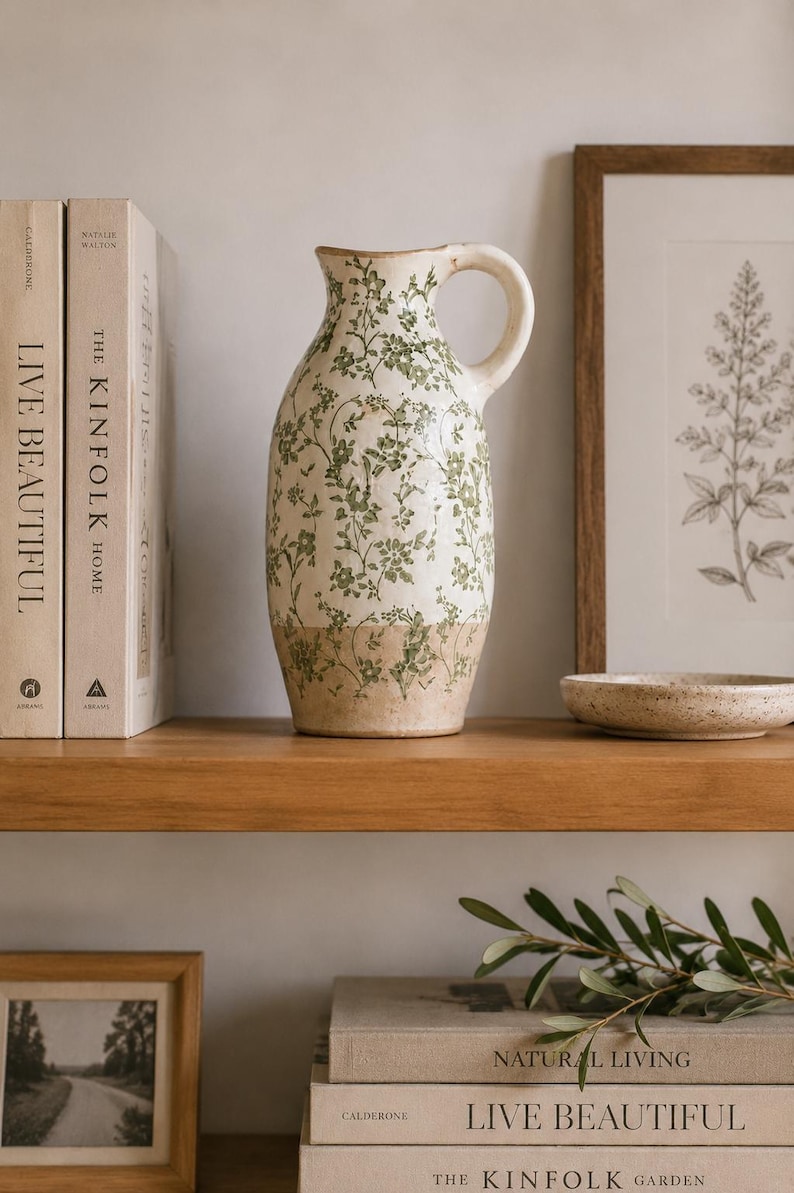 May include: A ceramic pitcher featuring a cream-colored base with a green floral vine pattern. The lower portion of the pitcher has a natural tan finish. It includes a curved handle on the right side and a slightly flared rim at the top opening.