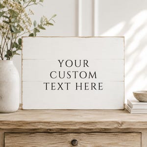 May include: A rectangular white wooden sign with horizontal plank detailing. The sign features the black printed text "YOUR CUSTOM TEXT HERE" centered on the front surface. The wood has a distressed finish with visible grain and texture.