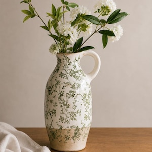May include: A ceramic pitcher with a cream-colored base and a green floral pattern featuring delicate vines and blossoms. The pitcher has a single side handle and contains a bouquet of white flowers with green leaves extending from the top opening.