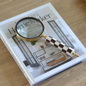 May include: A magnifying glass with a circular gold-toned metal frame and a cylindrical handle featuring a black and white checkered pattern. The magnifying glass is resting on top of a book titled Modern HomeMaker with a cover image of a room.