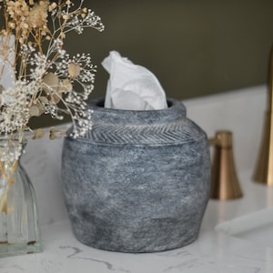 May include: A round, dark gray tissue holder with a textured, stone-like finish and a subtle herringbone pattern carved around the neck. The container has a wide opening at the top from which a single white tissue is protruding, showing its functional design.