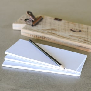 May include: A stack of white notepads with a black pencil on top. The notepads are on a light brown surface.