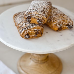 May include: Three chocolate croissants dusted with powdered sugar sit on a white marble cake stand with a wooden base. The pastries have a flaky, golden-brown crust and dark chocolate filling.