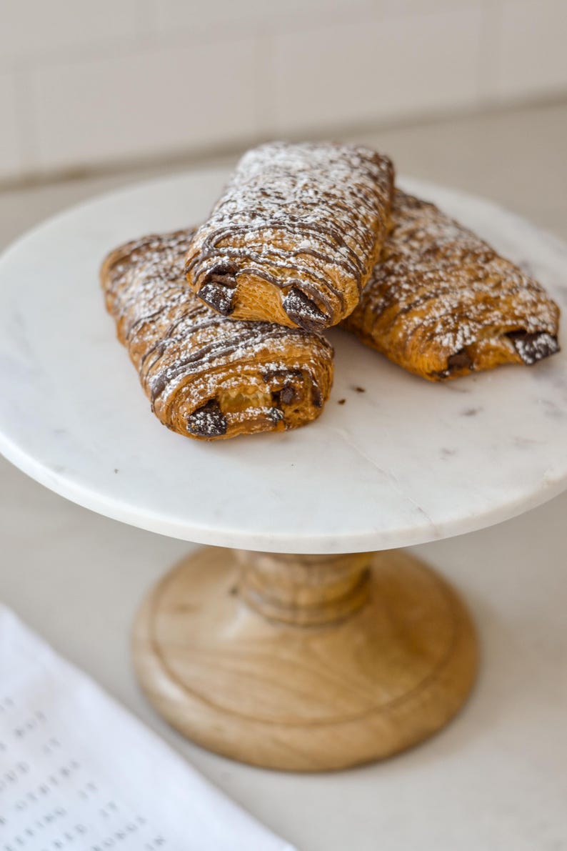 May include: Three chocolate croissants dusted with powdered sugar sit on a white marble cake stand with a wooden base. The pastries have a flaky, golden-brown crust and dark chocolate filling.